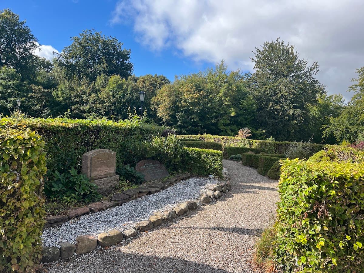 Gravel pathway through a garden with hedges and gravestones under a partly cloudy sky